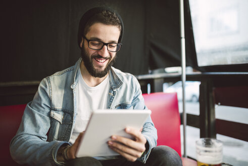 Porträt eines lächelnden jungen Mannes, der in einer Kneipe sitzt und ein Tablet benutzt - RAEF01812