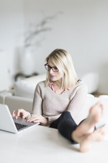 Blonde Frau sitzt mit hochgelegten Füßen am Tisch und benutzt einen Laptop - CHPF00379