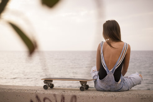 Rückenansicht einer Frau mit Skateboard, die auf einer Mauer vor dem Meer sitzt - SIPF01608