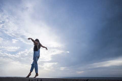 Frau mit ausgestreckten Armen balanciert auf einer Mauer vor dem Himmel, lizenzfreies Stockfoto