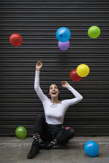 Lachende junge Frau, die auf dem Bürgersteig sitzt und fliegende Ballons beobachtet - KKAF00652