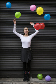 Glückliche junge Frau vor einem schwarzen Rollladen mit fliegenden Luftballons - KKAF00653