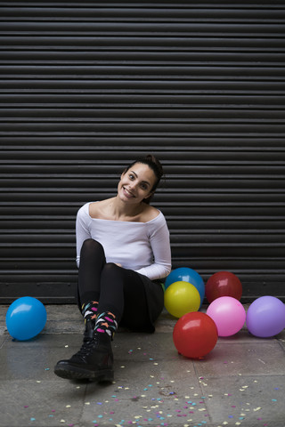 Lächelnde junge Frau sitzt mit Luftballons auf dem Bürgersteig, lizenzfreies Stockfoto