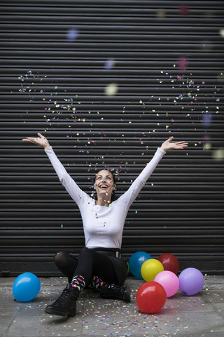 Happy young woman sitting on pavement with balloon throwing confetti stock photo