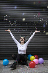 Glückliche junge Frau auf dem Bürgersteig sitzend mit Luftballon und Konfetti werfend - KKAF00656