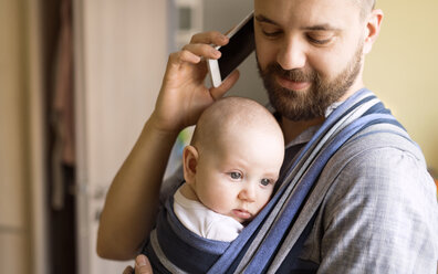 Vater mit Baby im Tragetuch zu Hause im Gespräch mit dem Handy - HAPF01415