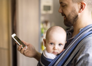 Vater mit Baby im Tragetuch zu Hause beim Telefonieren - HAPF01416