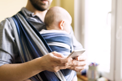 Vater mit Baby im Tragetuch zu Hause beim Telefonieren, lizenzfreies Stockfoto