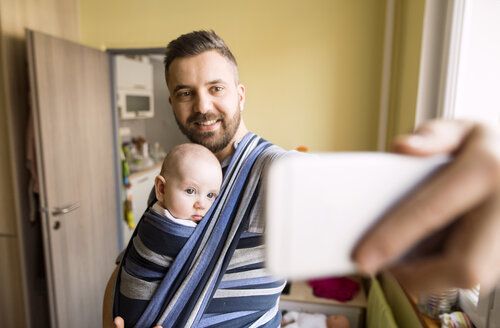 Vater mit Baby im Tragetuch macht zu Hause ein Selfie - HAPF01418