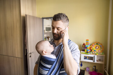 Vater mit Baby im Tragetuch zu Hause im Gespräch mit dem Handy - HAPF01419