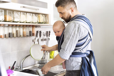 Father with baby son in sling at home doing the dishes - HAPF01420