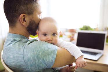 Vater hält Babysohn zu Hause mit Laptop auf dem Tisch - HAPF01434