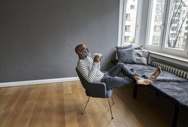 Älterer Mann mit Kopfhörern sitzt am Fenster und trinkt Kaffee - FMKF03748