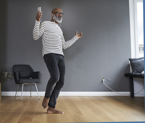 Älterer Mann tanzt allein zu Hause, hält Smartphone, lizenzfreies Stockfoto