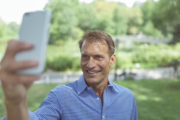 Porträt eines lächelnden Mannes, der ein Selfie mit seinem Smartphone in einem Park macht - BOYF00757