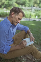 Mann mit Notizbuch sitzt auf einer Wiese in einem Park und schreibt etwas auf - BOYF00766