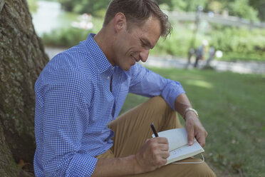 Mann mit Notizbuch sitzt auf einer Wiese in einem Park und schreibt etwas auf - BOYF00767