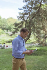 Mann mit Mini-Tablet, der auf einer Wiese in einem Park spazieren geht - BOYF00768