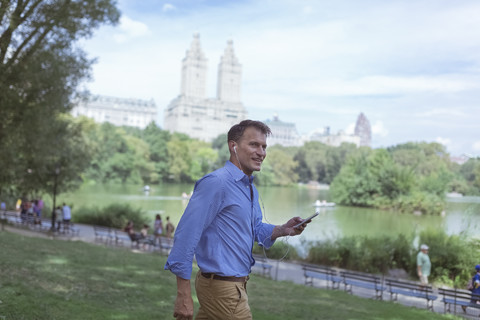 USA, Manhattan, Central Park, lächelnder Mann mit Mobiltelefon und Kopfhörern, lizenzfreies Stockfoto