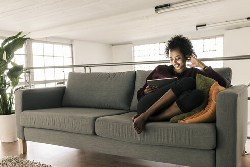 Smiling young woman sitting on couch using tablet - UUF10314