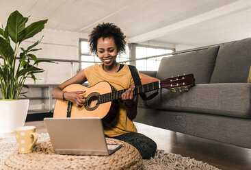 Lächelnde junge Frau, die zu Hause Gitarre spielt und auf ihren Laptop schaut - UUF10323