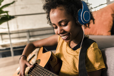Lächelnde junge Frau zu Hause mit Kopfhörern, die Gitarre spielt - UUF10327