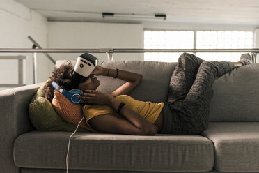 Junge Frau liegt mit Kopfhörern und VR-Brille auf der Couch - UUF10330