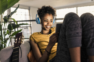 Smiling young woman on couch with headphones using tablet - UUF10334