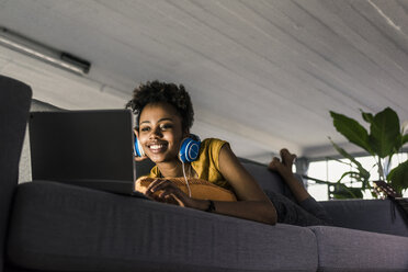 Smiling young woman with headphones lying on couch using laptop - UUF10338