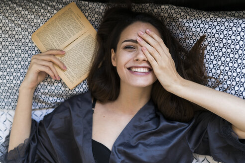 Portrait of smiling young woman lying on bed with a book covering one eye with her hand - KKAF00662