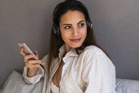 Porträt einer lächelnden jungen Frau mit Mobiltelefon, die zu Hause mit Kopfhörern Musik hört, lizenzfreies Stockfoto