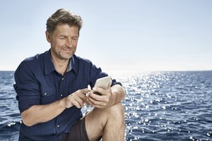Porträt eines lächelnden reifen Mannes vor dem Meer mit einem Mobiltelefon - PDF01109