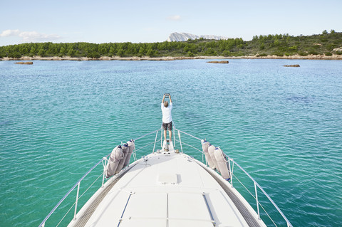 Rückansicht eines Mannes, der am Bug seiner Motoryacht steht und mit einem Tablet fotografiert, lizenzfreies Stockfoto