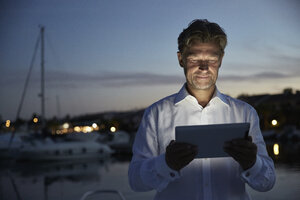 Porträt eines lächelnden reifen Mannes, der in der Abenddämmerung vor dem Hafen steht und auf eine Tafel schaut - PDF01148