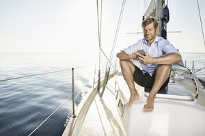 Lächelnder reifer Mann, der auf seinem Segelboot sitzt und ein Mobiltelefon benutzt - PDF01159