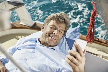 Portrait of smiling mature man lying on deck of his sailing boat looking at cell phone - PDF01191