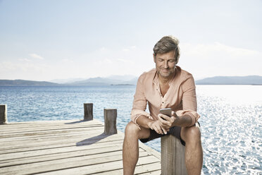 Smiling man sitting on jetty looking at cell phone - PDF01207