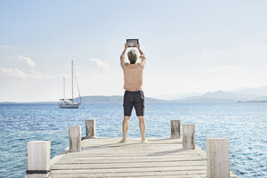 Rückansicht eines Mannes, der auf einem Steg steht und ein Selfie mit einem Tablet macht - PDF01209