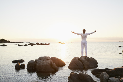 Rückenansicht eines Mannes, der auf einem Felsen steht und bei Sonnenuntergang auf das Meer blickt, lizenzfreies Stockfoto