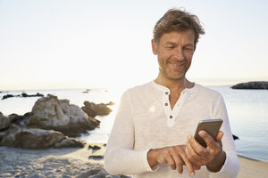 Porträt eines lächelnden Mannes, der am Strand ein Mobiltelefon benutzt - PDF01219