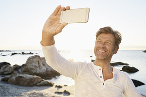 Porträt eines lächelnden Mannes, der mit seinem Handy ein Selfie am Strand macht - PDF01221