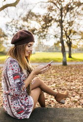 Lächelnde junge Frau sitzt auf einer Bank im herbstlichen Park und schaut auf ihr Handy - MGOF03202