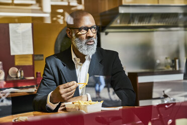 Mature businessman sitting in snack bar, eating French fries - FMKF03819