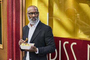 Älterer Geschäftsmann isst Pommes frites vor einer Snackbar - FMKF03820