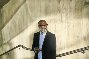 Mature businessman leaning against concrete wall, holding smartphone - FMKF03858