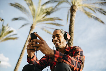 Spanien, Porträt eines lächelnden jungen Mannes, der mit Kopfhörern Musik hört und auf sein Mobiltelefon schaut - JRFF01289