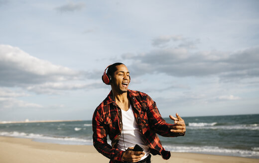 Portrait of young man on the beach singg and dancing while listening music with headphones - JRFF01294