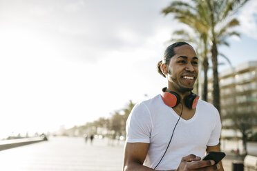 Spain, portrait of smiling young man with headphones and cell phone on beach promenade - JRFF01299