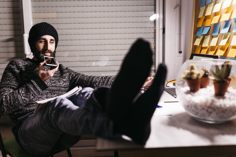 Man with feet up at his desk recording a voice note at night stock photo