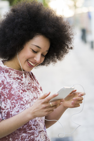 Porträt einer lächelnden jungen Frau mit Kopfhörern, die auf ihr Mobiltelefon schaut, lizenzfreies Stockfoto